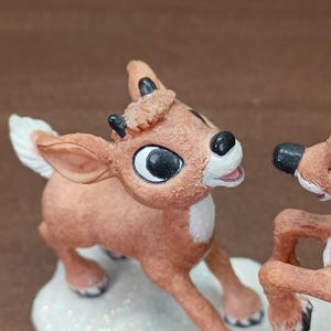 May include: A reindeer figurine with a red nose, black eyes, and small antlers. The reindeer is light brown with a white chest and stands on a snowy base. A Christmas decoration.