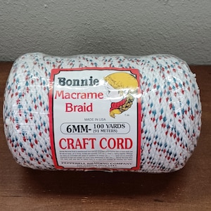 May include: A spool of Bonnie Macrame Braid craft cord wrapped in clear plastic. The cord is white with small red and blue flecks. The label reads Bonnie Macrame Braid, 6MM- 100 YARDS (91 METERS) CRAFT CORD, MADE IN USA, and Pepperell Braiding Company.