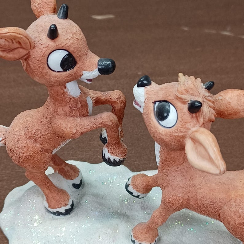 May include: Two brown reindeer figurines with large eyes and black antlers. One is standing on its hind legs, the other looking up. They are on a white, glittery base, suggesting a winter theme. The figurines are likely for decoration.