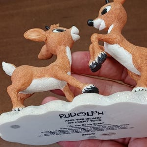 May include: A figurine set featuring two Rudolph the Red-Nosed Reindeer characters from "Rudolph and the Island of Misfit Toys." The figures are brown with white accents and stand on a snowy base. The base has text that reads "You Can Be My Buddy."