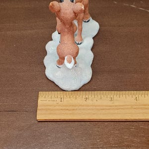 May include: A brown reindeer figurine with a white tail and black hooves stands on a white, glittery base. The figurine is approximately 5 cm tall. A wooden ruler with "MADE IN U.S.A." is in the foreground.