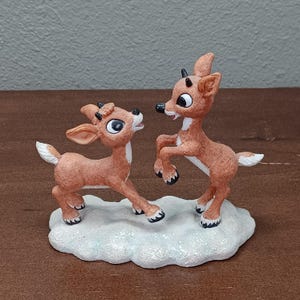 May include: A festive figurine of two reindeer in a playful pose. The reindeer are light brown with white accents and black hooves and antlers. They stand on a white, glittery base, creating a festive holiday scene.