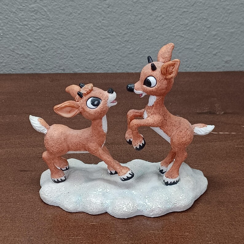 May include: A festive figurine of two reindeer in a playful pose. The reindeer are light brown with white accents and black hooves and antlers. They stand on a white, glittery base, creating a festive holiday scene.
