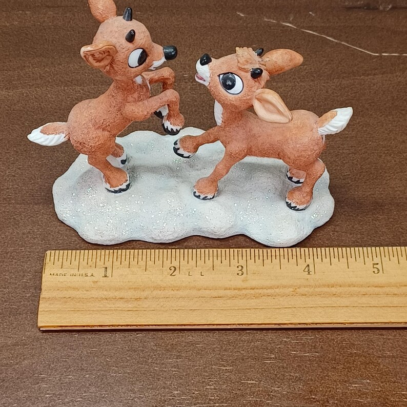 May include: A Christmas figurine of two reindeer in a playful pose on a snow-covered base. The reindeer are brown with white accents and black hooves. The base is white with glitter. A wooden ruler with inch measurements is in the foreground.