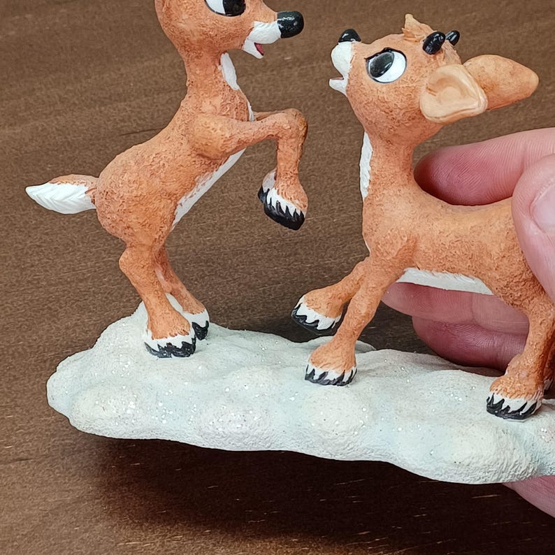 May include: A figurine of two reindeer in a playful pose. The reindeer are brown with white accents and black hooves. They stand on a white, snow-like base. The figurine is likely a Christmas decoration.