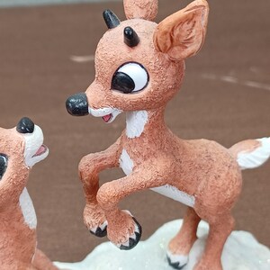 May include: A detailed figurine of a reindeer with a brown body, white patches, and black antlers. The reindeer is posed mid-jump, with a white tail and black hooves. The figurine is on a snowy base, with a second reindeer visible.