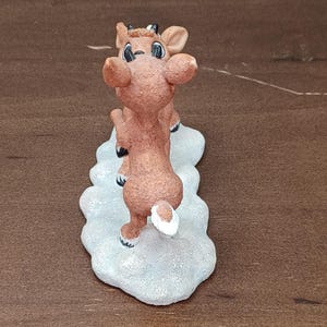May include: A brown reindeer figurine with a white tail and black eyes stands on a snow-covered base. The reindeer is in a playful pose, with its front legs raised. The figurine is a festive holiday decoration.