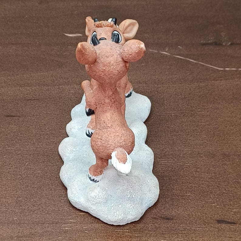 May include: A brown reindeer figurine with a white tail and black eyes stands on a snow-covered base. The reindeer is in a playful pose, with its front legs raised. The figurine is a festive holiday decoration.