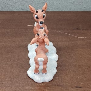 May include: A brown reindeer figurine with a red nose and large eyes, standing on a white base. The figurine features two reindeer, one standing on top of the other. The reindeer are in a playful pose, with one looking up at the other.