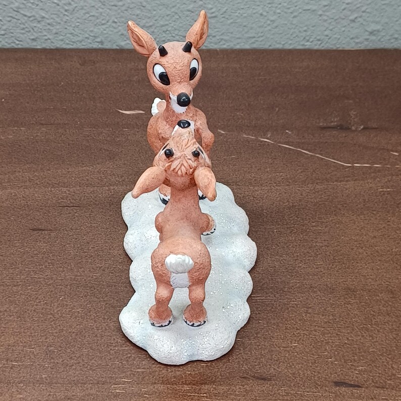May include: A brown reindeer figurine with a red nose and large eyes, standing on a white base. The figurine features two reindeer, one standing on top of the other. The reindeer are in a playful pose, with one looking up at the other.
