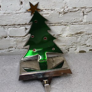 May include: A Christmas stocking holder shaped like a green Christmas tree with a gold star on top. The tree has red accents and is made of shiny metal. The holder has a silver hook for hanging.