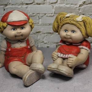 May include: Two ceramic figurines of children, one wearing a red and white baseball cap and overalls, the other wearing a red dress with white trim and pigtails.