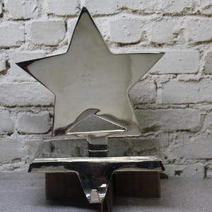 May include: A silver-colored metal stocking holder shaped like a star. The star is mounted on a rectangular base with a hook for hanging. The background is a white brick wall.
