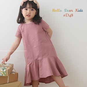 May include: A young girl wearing a pink linen dress with a ruffled hem and a floral headband. She is standing on a wooden floor with a stack of wrapped gifts in front of her. The text "Hello Dear Kids #D48" is written in colorful letters on a white background.