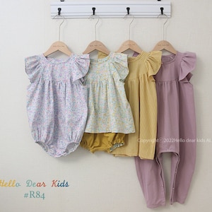 May include: Four children's clothing items hanging on a white rack. The items are a floral print romper, a floral print dress with a yellow waistband, a yellow and white striped dress, and a solid purple romper. The text "Hello Dear Kids #R84" is at the bottom of the image.