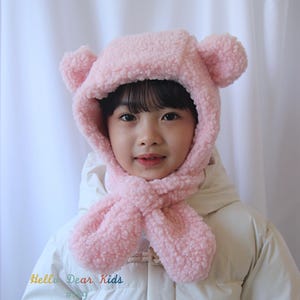 May include: A child wearing a pink, fluffy bear hat with matching scarf. The hat has two round ears on top. The child is wearing a white winter coat. The text "Hello Dear Kids" is visible in the lower left corner.