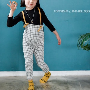 May include: A young girl wearing a black and white checkered jumpsuit with a brown leather strap. She is wearing a yellow and black striped headband that says "Hello" and is holding a small yellow object in her hand. She is standing in front of a blue wall with a potted plant in the corner.