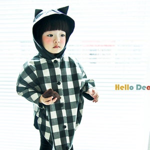 May include: A young child wearing a black and white checkered raincoat with a black cat ear hood. The child is holding a chocolate chip cookie and is standing in front of a window with white blinds. The text "Hello Dear Kids #06" is visible in the image.