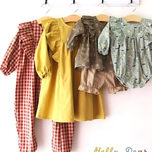 May include: Five children's clothing items hanging on a white rack. The items include a red and white gingham jumpsuit, a yellow dress with ruffles, a green and brown patterned blouse with matching trousers, and a green and white patterned onesie. The rack has the word "hello" written on it and the hashtag "#R52".
