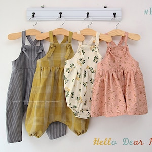 May include: Four different styles of children's dungarees, hanging on wooden hangers. The dungarees are made of different fabrics, including a grey striped fabric, a yellow checked fabric, a white fabric with a floral pattern, and a pink fabric with a floral pattern. The dungarees all have a criss-cross strap design at the back and a bow tie detail at the neck. The text "#R57" is visible in the top right corner of the image. The text "Hello Dear Kids" is visible in the bottom right corner of the image.