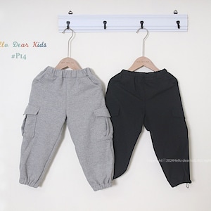 May include: Two pairs of children's cargo pants hanging on a white rack. One pair is gray, the other is black. Both have elastic waistbands and pockets on the sides. The text "Hello Dear Kids #P14" is visible.