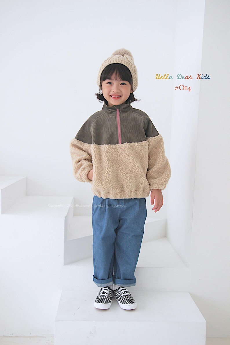 Pu&ograve; includere: Un bambino indossa una giacca di pile marrone e beige con una cerniera rosa, jeans blu e un berretto di lana bianco con un pon pon. Il testo "Hello Dear Kids #014" &egrave; nell'angolo in alto a destra dell'immagine.