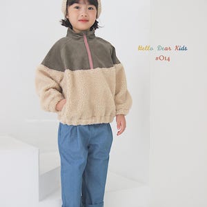 Pu&ograve; includere: Una persona giovane indossa una giacca di pile verde e beige con una cerniera rosa, jeans blu e scarpe da ginnastica a scacchi. La persona indossa anche un berretto di lana beige. Il testo "Hello Dear Kids #014" &egrave; visibile nell'immagine.