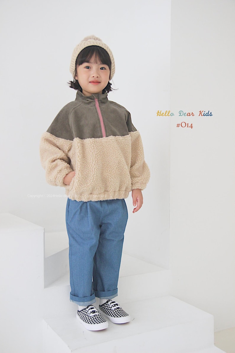 Pu&ograve; includere: Una persona giovane indossa una giacca di pile verde e beige con una cerniera rosa, jeans blu e scarpe da ginnastica a scacchi. La persona indossa anche un berretto di lana beige. Il testo "Hello Dear Kids #014" &egrave; visibile nell'immagine.