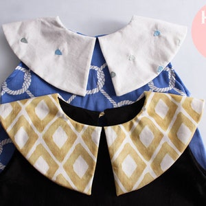 Puede incluir: Dos cuellos de tela diferentes, uno blanco con puntos bordados azules y otro amarillo y blanco con un patrón geométrico. Los cuellos están encima de dos camisas de colores diferentes, una azul y otra negra.