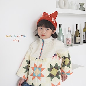 Pu&ograve; includere: Una giovane persona indossa una giacca trapuntata bianca e multicolore con un berretto rosso. La giacca ha una cerniera e un colletto alto. Il berretto ha un logo rosso e bianco che dice "Dear Kids". La persona indossa jeans blu e si trova davanti a un muro bianco.