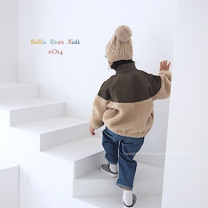 Pu&ograve; includere: Un bambino indossa una giacca di pile marrone e beige con colletto in velluto a coste marrone, jeans blu e un berretto di lana beige con un pon pon. Il bambino sta salendo una scala bianca. Il testo "Hello Dear Kids #014" &egrave; visibile nell'angolo in alto a sinistra dell'immagine.
