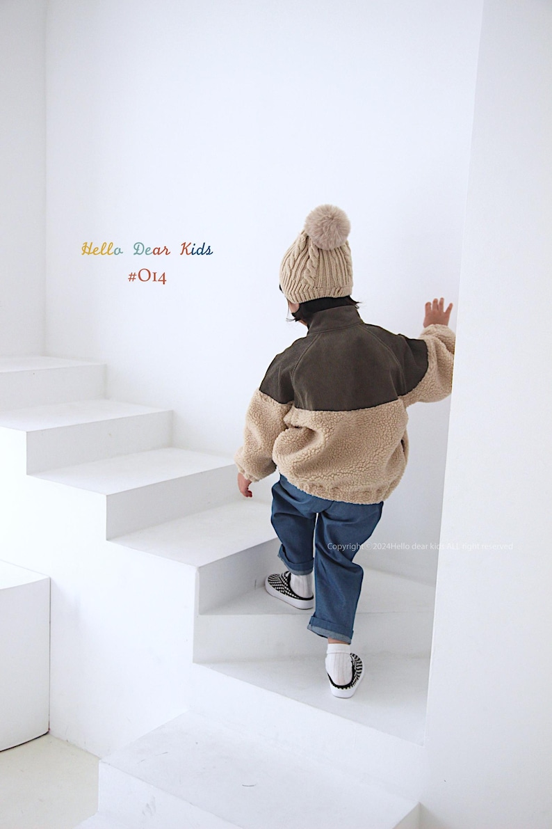 Pu&ograve; includere: Un bambino indossa una giacca di pile marrone e beige con colletto in velluto a coste marrone, jeans blu e un berretto di lana beige con un pon pon. Il bambino sta salendo una scala bianca. Il testo "Hello Dear Kids #014" &egrave; visibile nell'angolo in alto a sinistra dell'immagine.