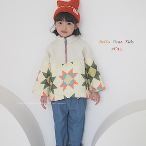 Pu&ograve; includere: Un bambino indossa una giacca trapuntata bianca e gialla con un motivo a stelle, jeans blu, scarpe marroni e un cappello di lana arancione con un logo. Il testo "Hello Dear Kids #014" &egrave; visibile nell'immagine.