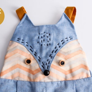 May include: A blue and orange baby onesie with a fox face design. The fox has black eyes and a black nose. The onesie has yellow straps.