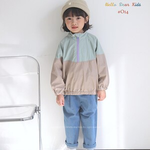 Pu&ograve; includere: Una giovane persona indossa una giacca blu chiaro e beige con una cerniera viola, jeans blu e un berretto bianco. La giacca ha un design a blocchi di colore. La persona &egrave; in piedi davanti a un muro bianco con una sedia trasparente a sinistra. Il testo "Hello Dear Kids #014" &egrave; nell'angolo in alto a destra dell'immagine.