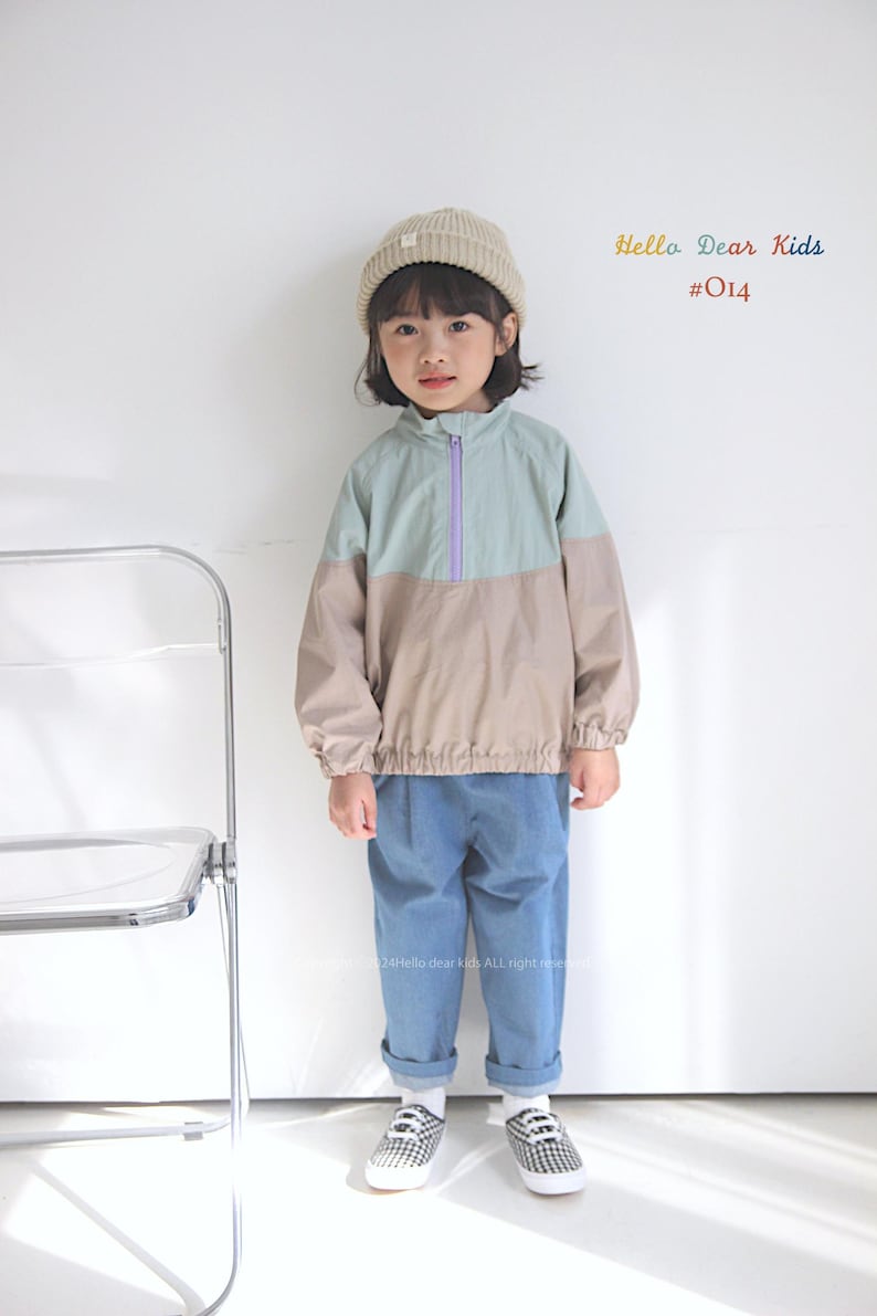 Pu&ograve; includere: Una giovane persona indossa una giacca blu chiaro e beige con una cerniera viola, jeans blu e un berretto bianco. La giacca ha un design a blocchi di colore. La persona &egrave; in piedi davanti a un muro bianco con una sedia trasparente a sinistra. Il testo "Hello Dear Kids #014" &egrave; nell'angolo in alto a destra dell'immagine.