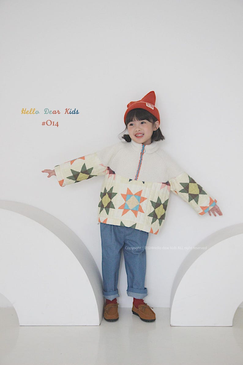 Pu&ograve; includere: Una persona giovane che indossa una giacca trapuntata bianca e multicolore con un berretto rosso. La giacca ha un motivo a stelle e il berretto ha un piccolo logo. La persona indossa jeans blu e scarpe marroni. Il testo "Hello Dear Kids #014" &egrave; visibile nell'angolo in alto a sinistra dell'immagine.