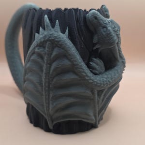May include: A dark gray ceramic mug with a dragon design. The mug features a dragon wrapped around the body, with a handle and a textured exterior. The dragon has detailed scales and a menacing expression. The mug is approximately 5 inches tall.