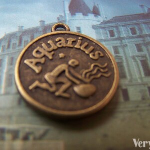 May include: A bronze-colored metal charm with the word "Aquarius" and a stylized image of the astrological sign.