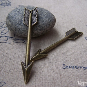 May include: Two antique gold arrow charms with a pointed tip and a decorative arrowhead. The charms have a hole at the top for attaching to jewellery or other crafts.