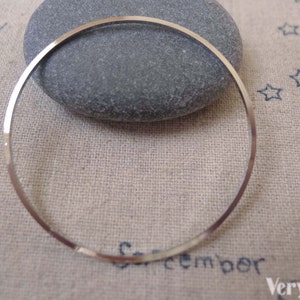 Può includere: Un cerchio sottile in metallo color argento, possibilmente un componente di gioielleria, poggia su una pietra grigia. Il cerchio è liscio e riflettente, con una leggera curvatura. Lo sfondo è un tessuto di colore chiaro.