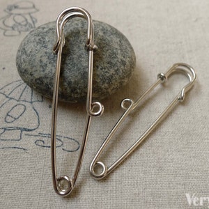 Op de afbeelding: Twee zilveren veiligheidsspelden, waarvan één groter is dan de andere, liggen op een beige stoffen oppervlak. Beide spelden zijn open en klaar voor gebruik.