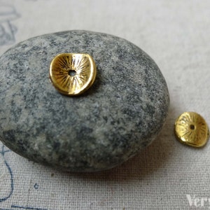 May include: Two small, gold-coloured metal bead caps with a textured, sunburst design. One bead cap is on a grey stone, and the other is next to it. The bead caps have a small hole in the centre.