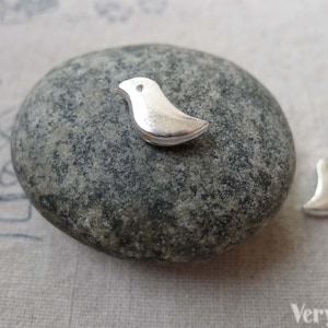 Könnte beinhalten: Zwei silberne Vogel-Anhänger, einer auf einem grauen Stein und einer daneben. Die Anhänger sind schlicht und minimalistisch im Design.