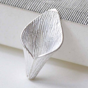 May include: A silver-coloured metal flower charm in the shape of a calla lily. The charm has a textured surface and a delicate, curved design.