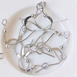 May include: A close-up of a pile of silver earring hooks with small round balls at the top. The hooks are made of metal and are designed to be used with earrings.
