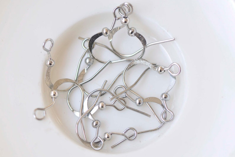 May include: A close-up of a pile of silver earring hooks with small round balls at the top. The hooks are made of metal and are designed to be used with earrings.