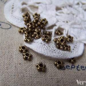 May include: A close-up of a group of small, antique gold-toned metal beads with a starburst design. The beads are scattered on a white lace doily and a brown fabric surface.