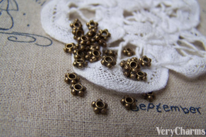 May include: A close-up of a group of small, antique gold-toned metal beads with a starburst design. The beads are scattered on a white lace doily and a brown fabric surface.