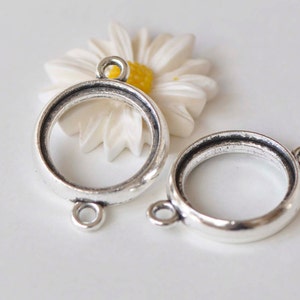 Op de afbeelding: Twee zilverkleurige metalen bezel-instellingen voor het maken van sieraden. Elke instelling heeft een ronde opening en twee springringen.